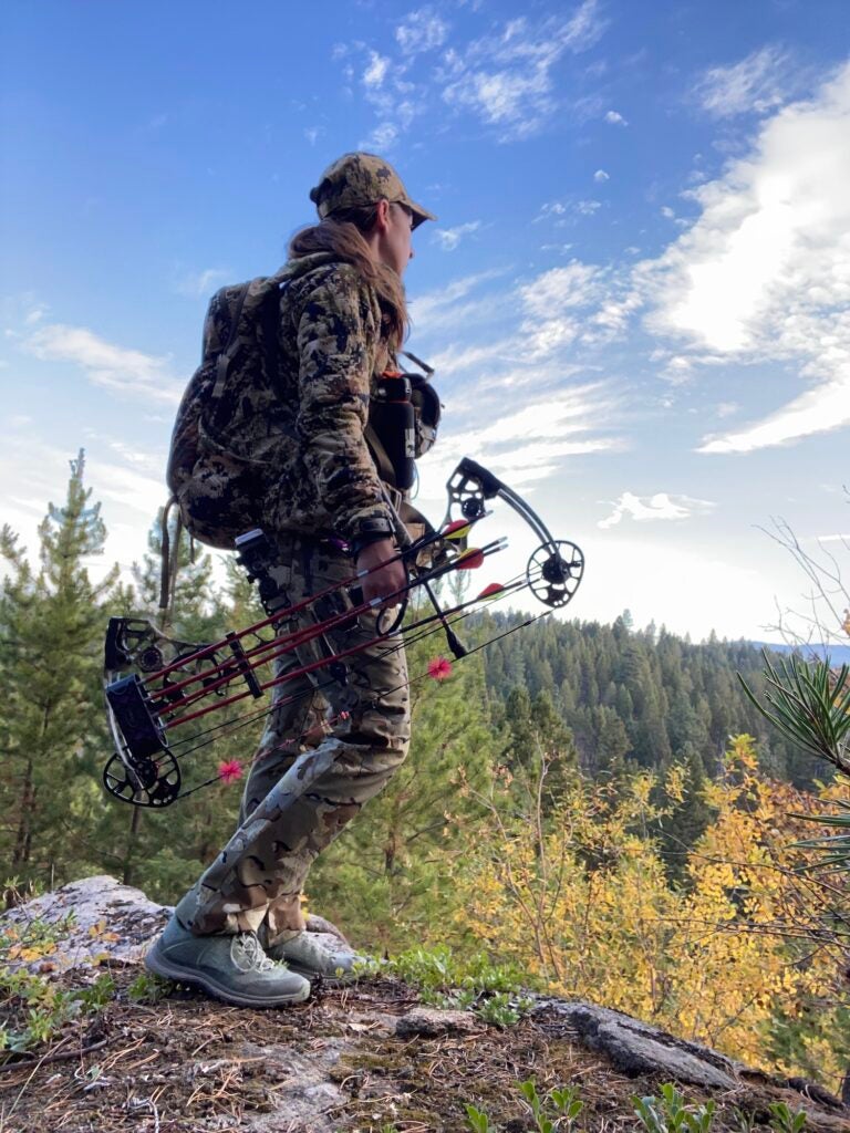 A woman in all camo standing on a rock holding a bow