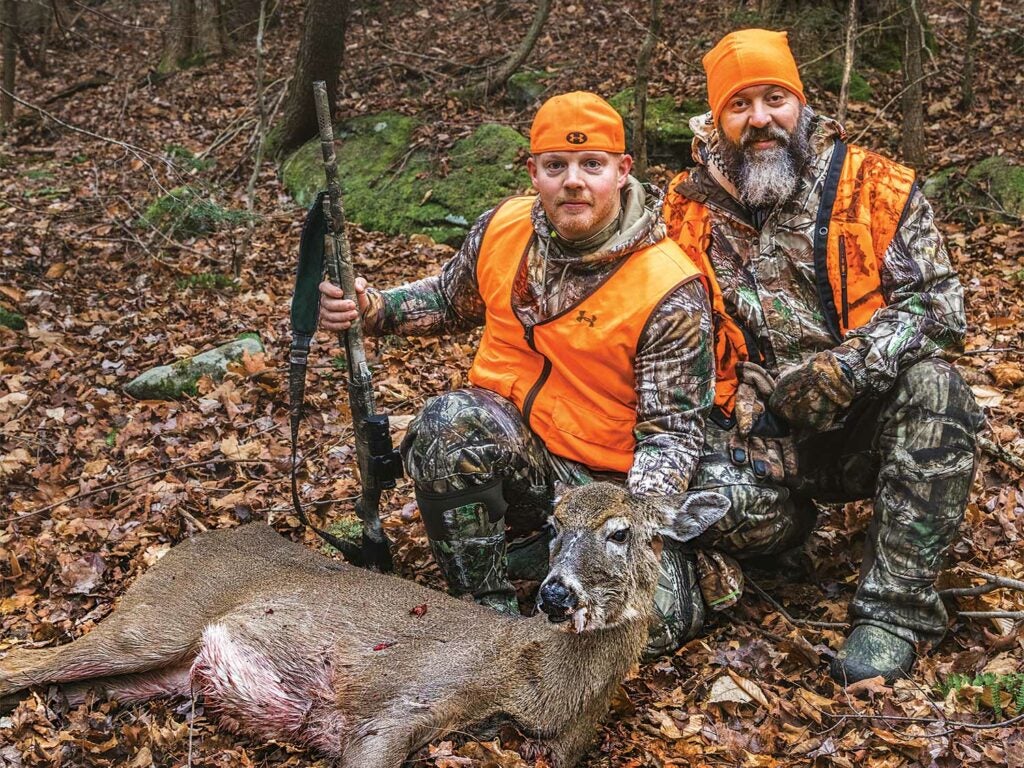 two hunters in orange kneeling in the leaves behind a dead doe