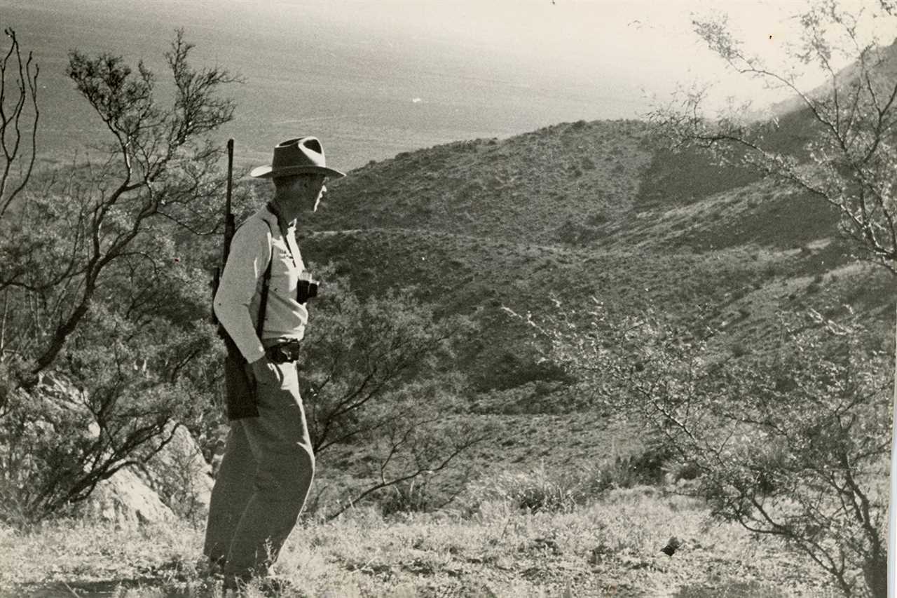 Hunter with gun and camera looks out onto arid vista.