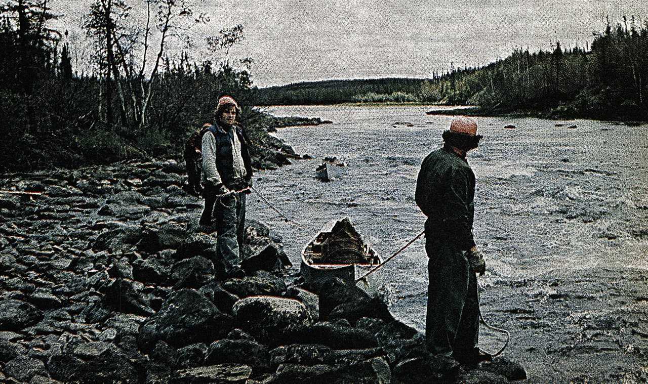 two canoeists pull canoe along in river from the rocky bank