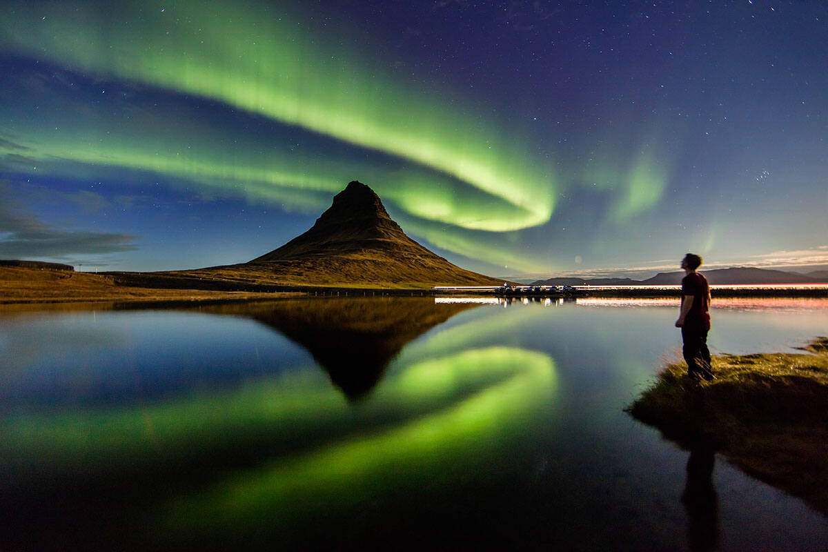 A man standing by a lake - there is a mountain on the other side of the lake and the Northern Lights are its backdrop