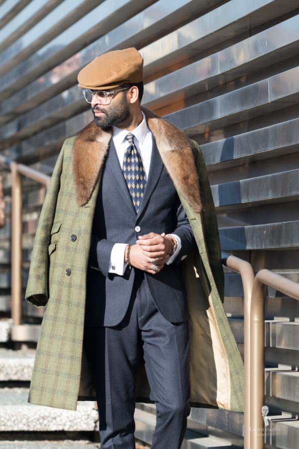 Green triple checked overcoat with fur collar, dark suit, 90s tie and white shirt with flat cap and clear glasses