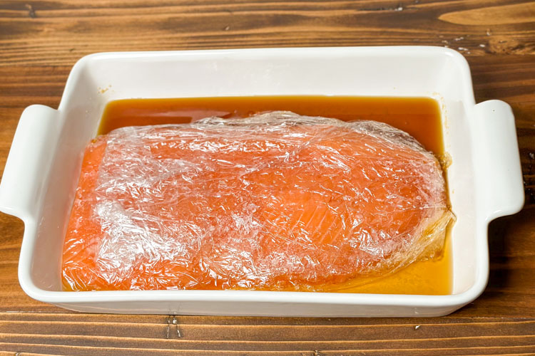 salmon filet wrapped in plastic wrap, in a white dish with juices