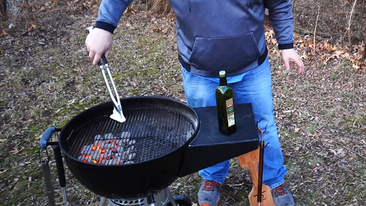 a person holding tongs with oil covered paper towel seasoning the charcoal grill grates