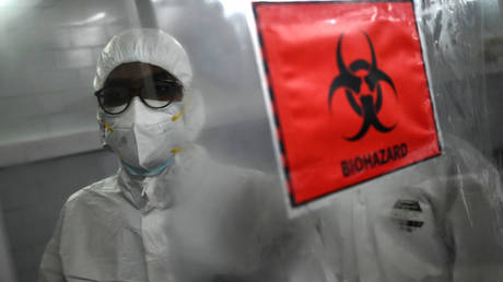 Technicians wearing personal protective equipment suits stand behind a biohazard sign.