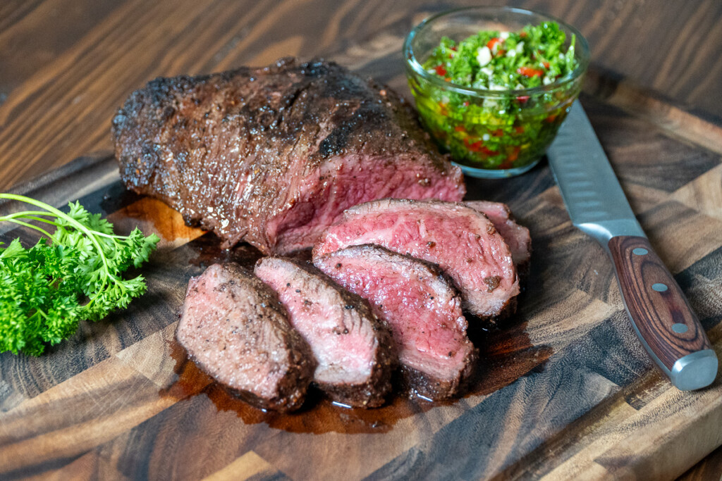 tri-tip sliced on a wooden board with a bowl of chimichurri