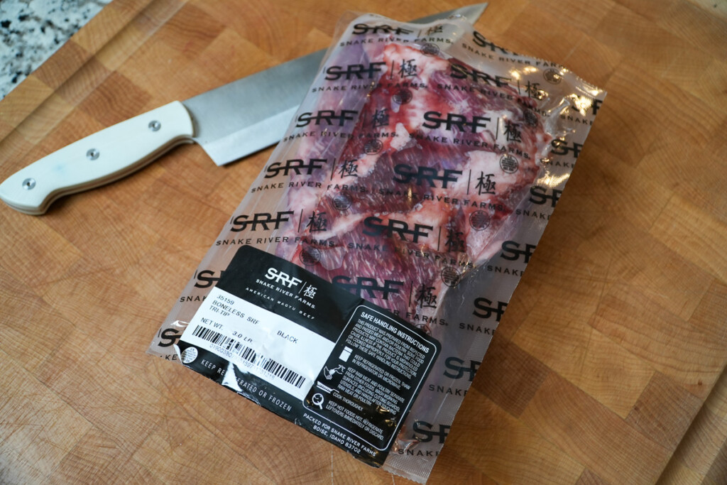 a plastic wrapped piece of tri-tip on a wooden chopping board with a knife in the background