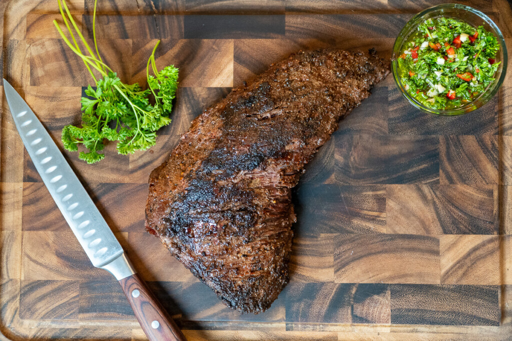 a piece of cooked tri-tip, a knife and some parslet on a wooden chopping board