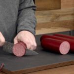 a man holding a knife cutting into a smoked summer sausage
