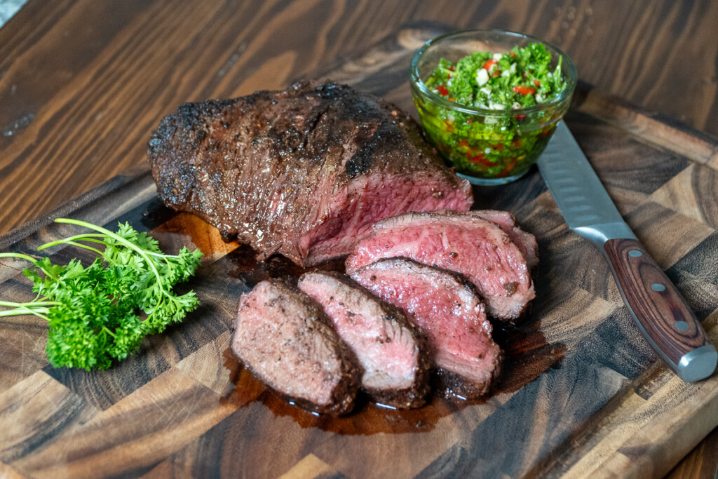 A steak sliced on a wooden board with a glass bowl of chimichurri
