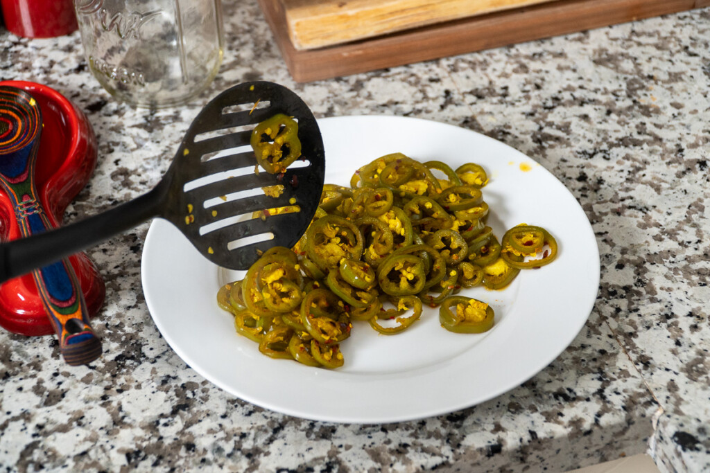 a white plate with cooked jalapenos on it and a slotted black spoon