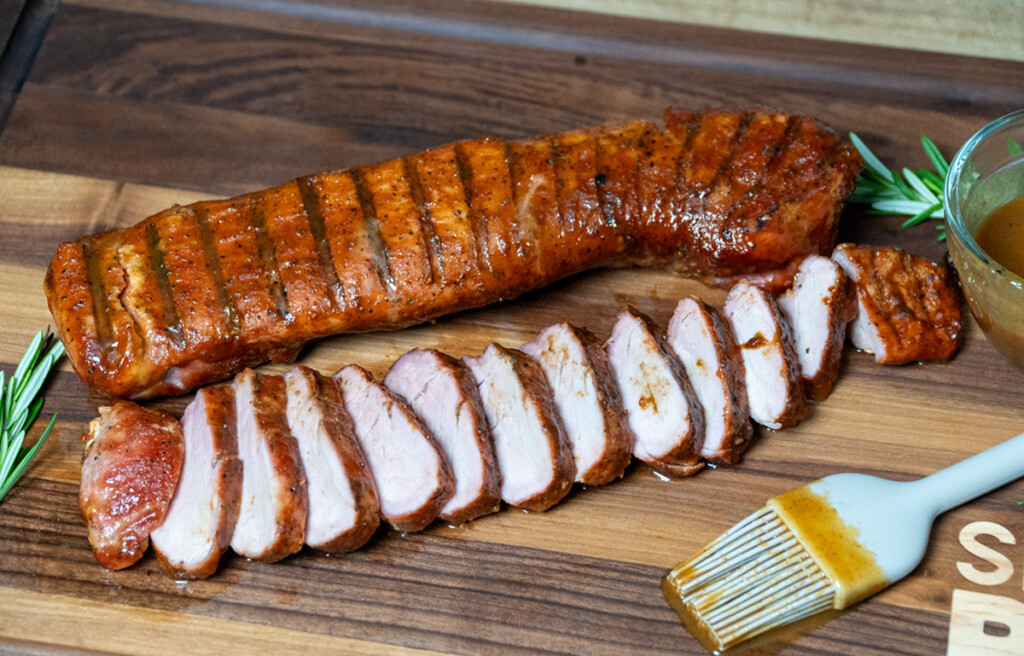 smoked pork tenderloin, sliced, on wooden board with basting brush beside