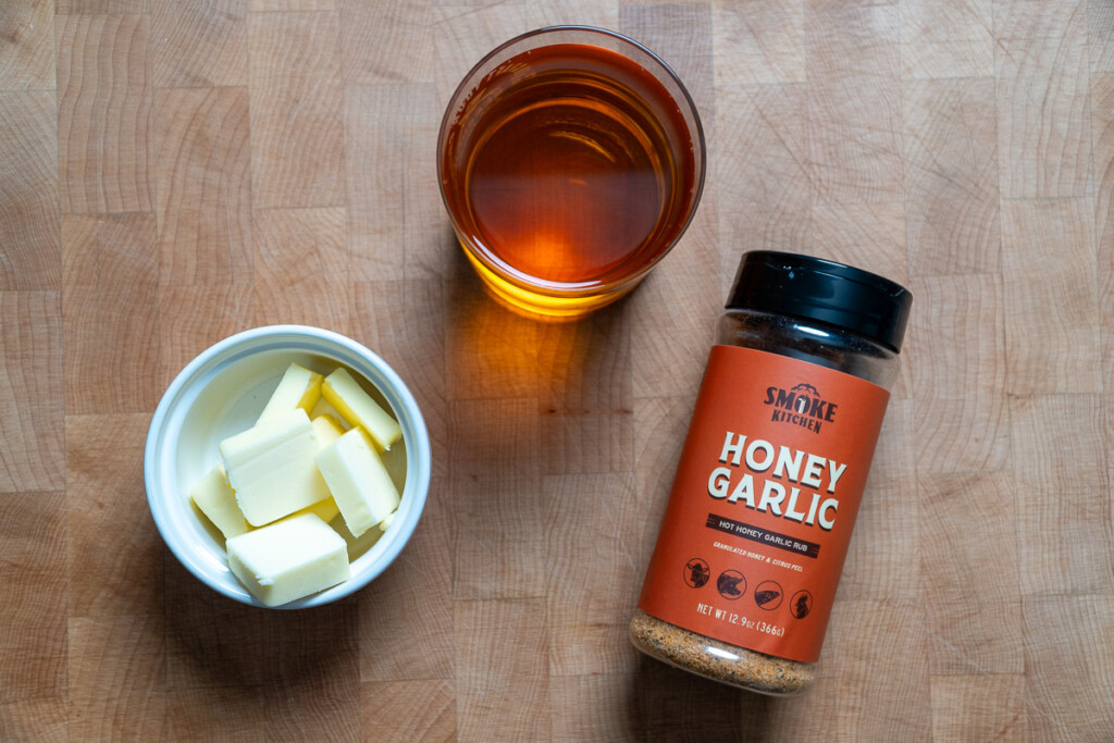 a white bowl of butter, glass of apple juice, jar of bbq rub on a wooden chopping board