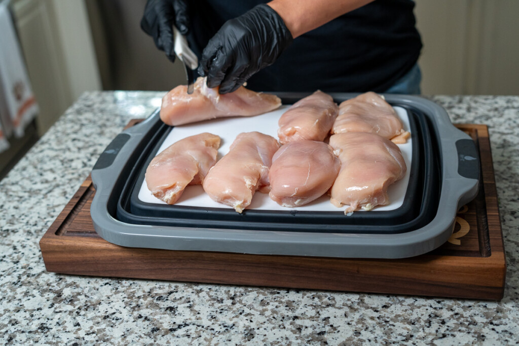 gloved hands holding a raw chicken breast removing fat