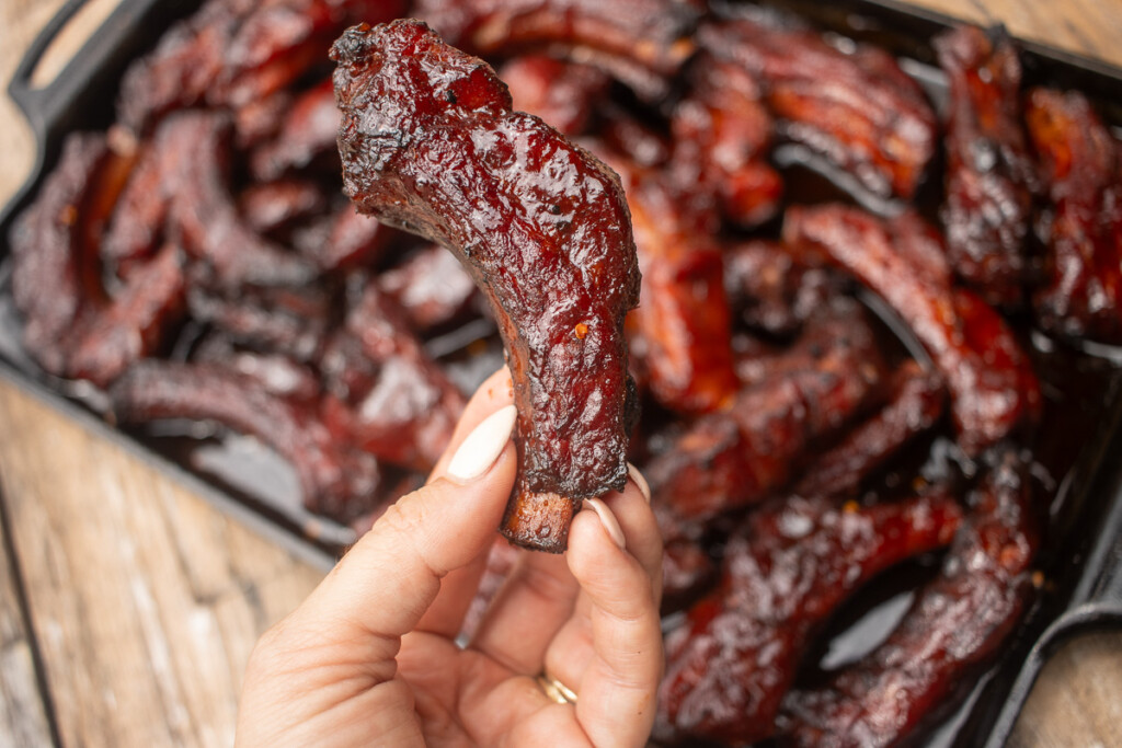 a hand holding a single party rib with a tray of ribs in the background