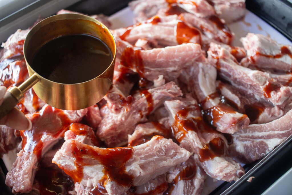 a measuring cup pouring BBQ sauce over raw ribs