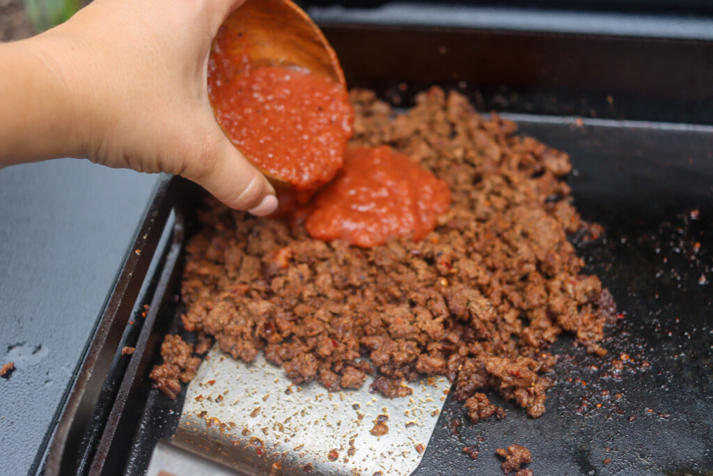 ground beef cooking on the griddle and a hand pour salsa from a cup onto it