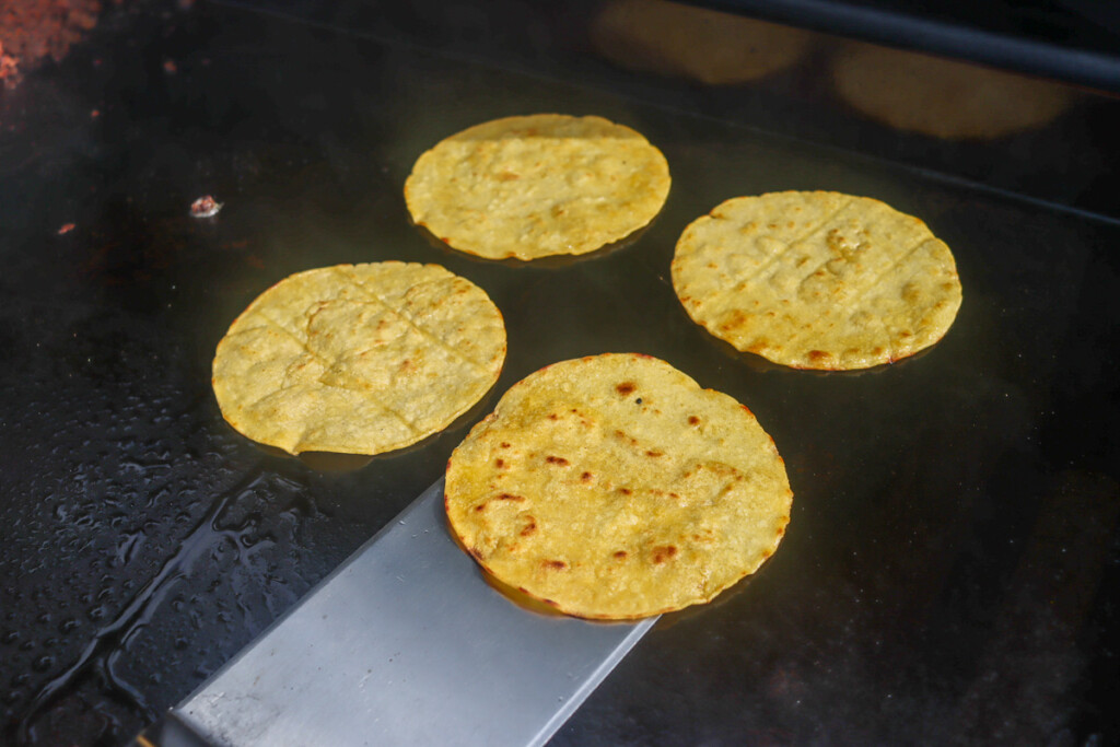 corn tortillas cooking on a griddle, one being turned with a spatula