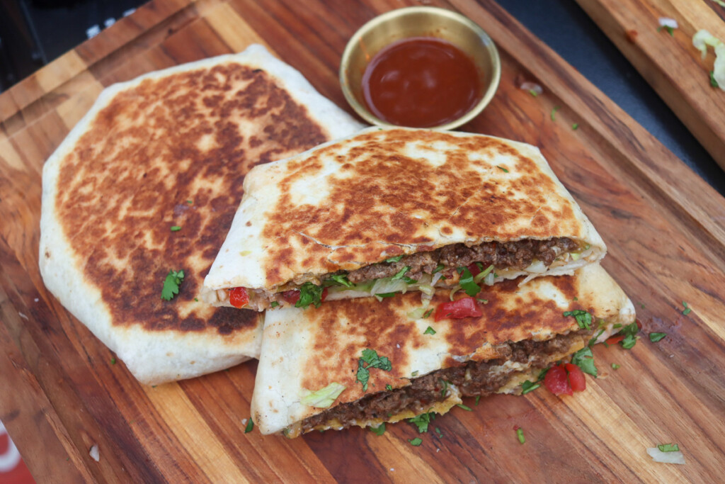 a crunchwrap cut in half and a whole cruchwrap with a bowl of hot sauce on a wooden board