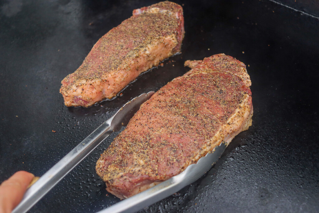 tongs placing a raw seasoned steak on the griddle