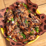 0-400 chicken wings garnished with sesame seeds and sliced green onions, served on brown parchment paper with barbecue-themed graphics, placed in a yellow basket on a wooden table.