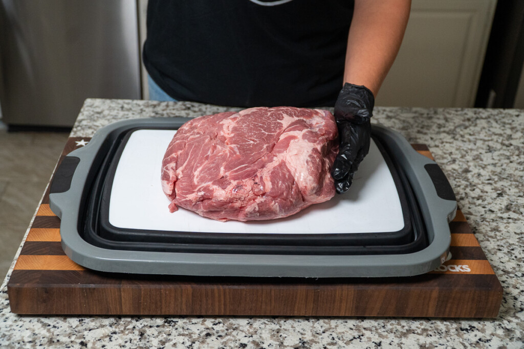 a raw piece of pork butt on a white board being with a gloved black hand on the right of it