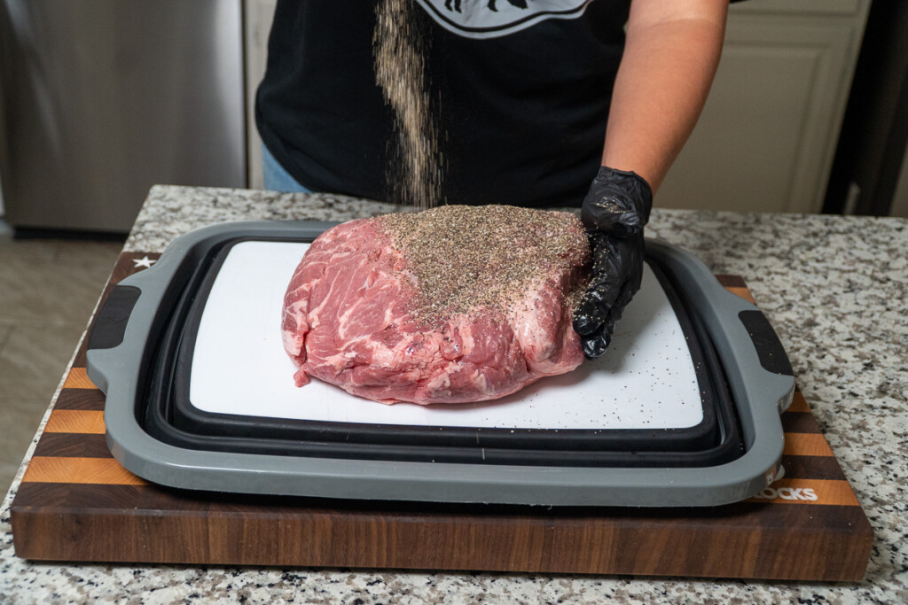 a person sprinkling BBQ rub over the raw pork butt