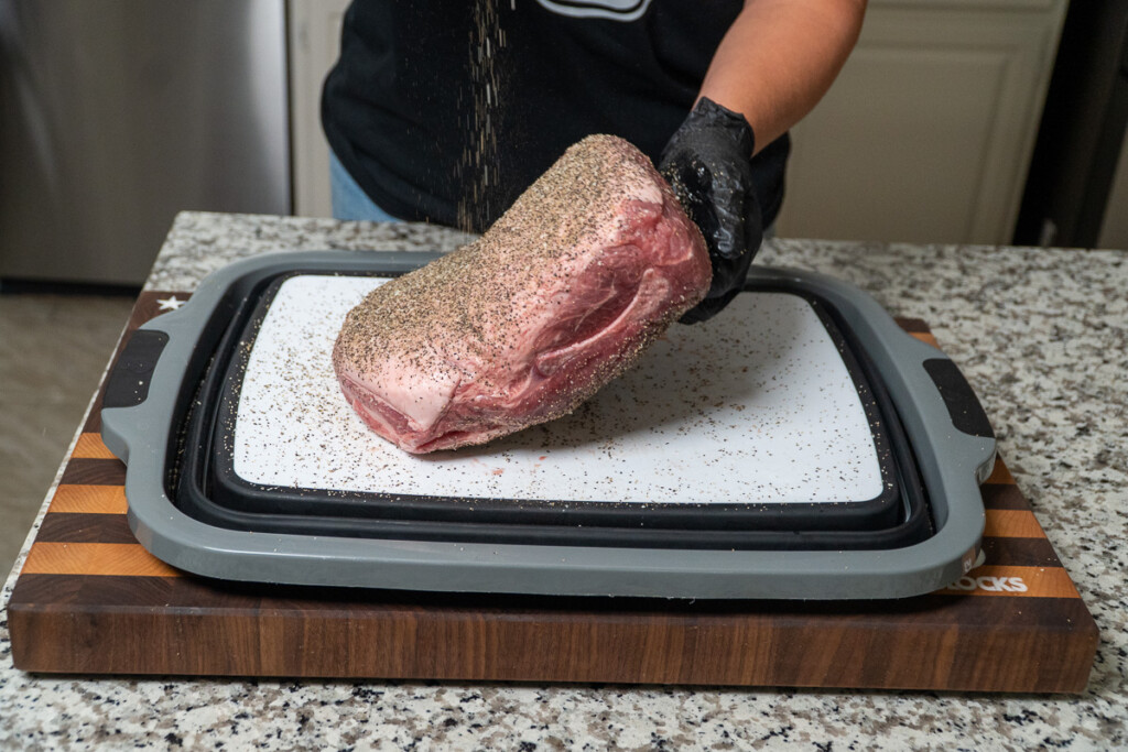 a person sprinkling BBQ rub over a raw pork butt