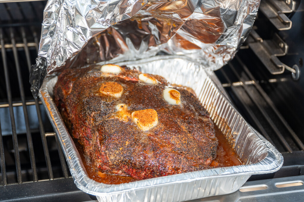 a pork butt in the smoker with pats of butter on top being covered with aluminum foil