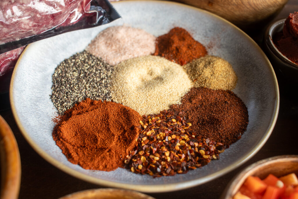 A ceramic plate displaying an assortment of chili seasoning spices, including ground black pepper, salt, garlic powder, onion powder, smoked paprika, chili powder, red pepper flakes, and cumin powder, arranged in small piles.