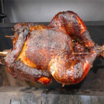 The image shows a well-cooked, crispy, and browned turkey mounted on a rod in a roasting oven. This process suggests an effective rotispliance technique, possibly involving a smoker given the rich coloring and texture of the turkey's skin.