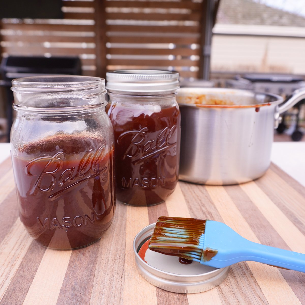 homemade bbq sauce in mason jars on a cutting board with a saucepan and dirty blue silicone brush.