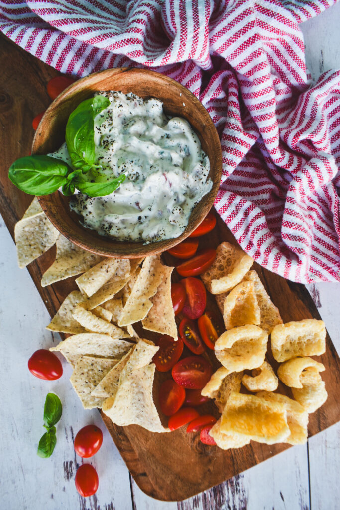 white blt dip in wooden bowl paired with tortilla chips, tomatoes and fresh basil on a white backdrop.