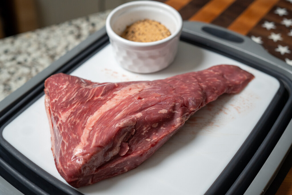 a raw pice of tri-tip on a white board with a bowl of seasoning beside it