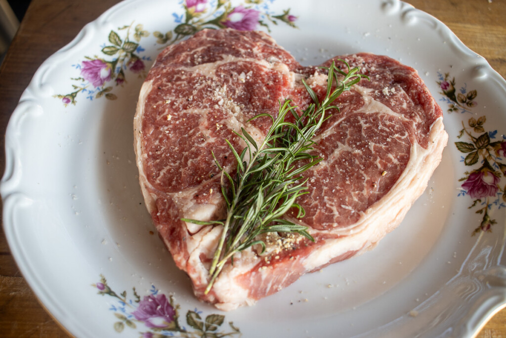 a raw heart-shape ribeye steak with herbs on it