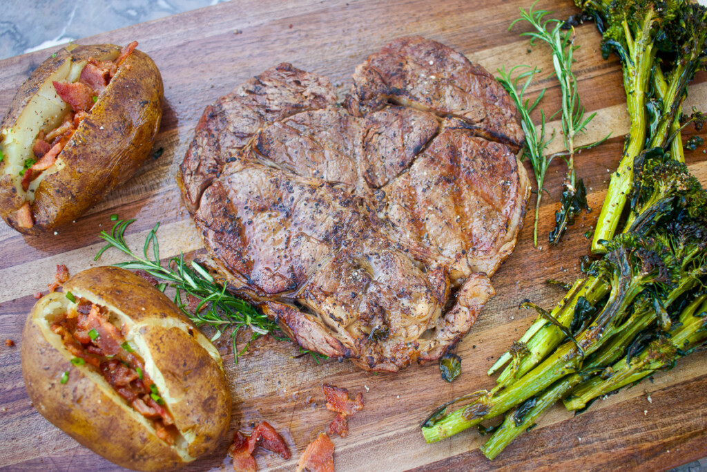 heart-shaped ribeye stead, baked potatoes and broccalli on a wooden board