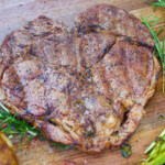 A heart-shaped ribeye steak with a beautifully seared crust sits on a wooden cutting board, surrounded by fresh rosemary, roasted asparagus, and a baked potato. The steak is seasoned with salt and pepper, glistening with juices, making it a perfect centerpiece for a special meal.