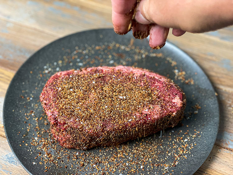 A close-up of a thick-cut steak on a dark plate, generously coated with a coffee-based dry rub. A hand is seen sprinkling additional rub over the meat, highlighting the rich, textured seasoning.