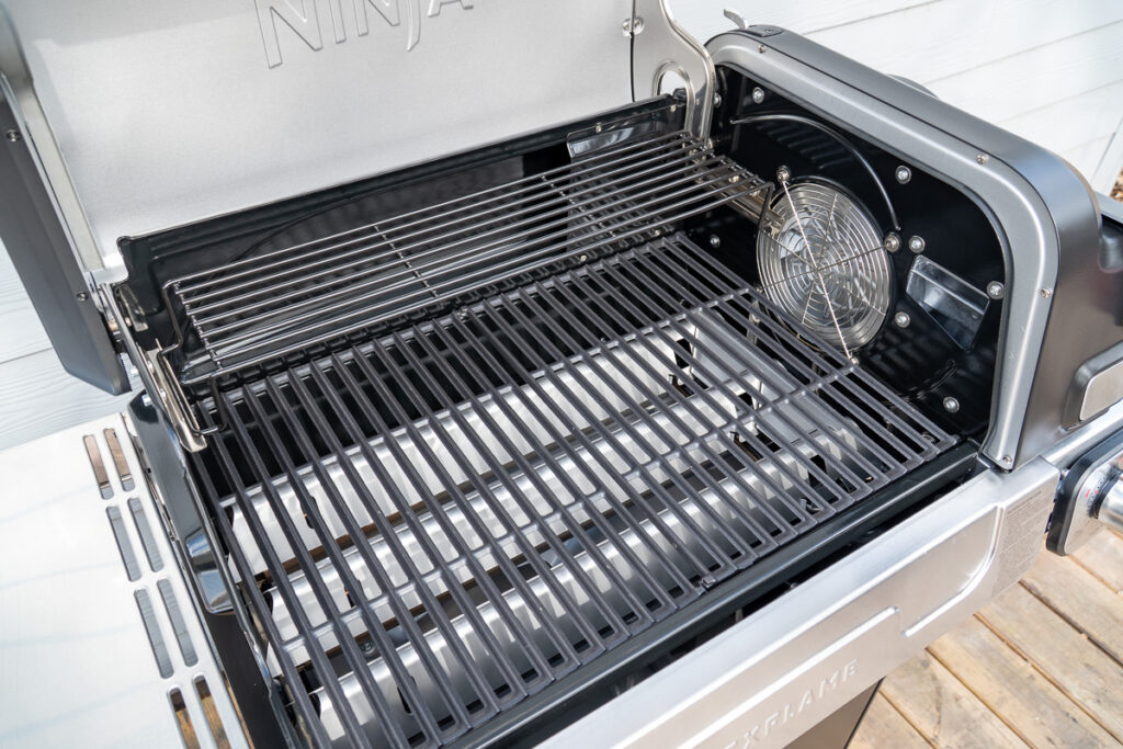 Interior of the Ninja FlexFlame grill with open lid, showcasing enameled cast iron grates, upper warming rack, and the integrated Cyclonic Heat IQ fan on the right side.