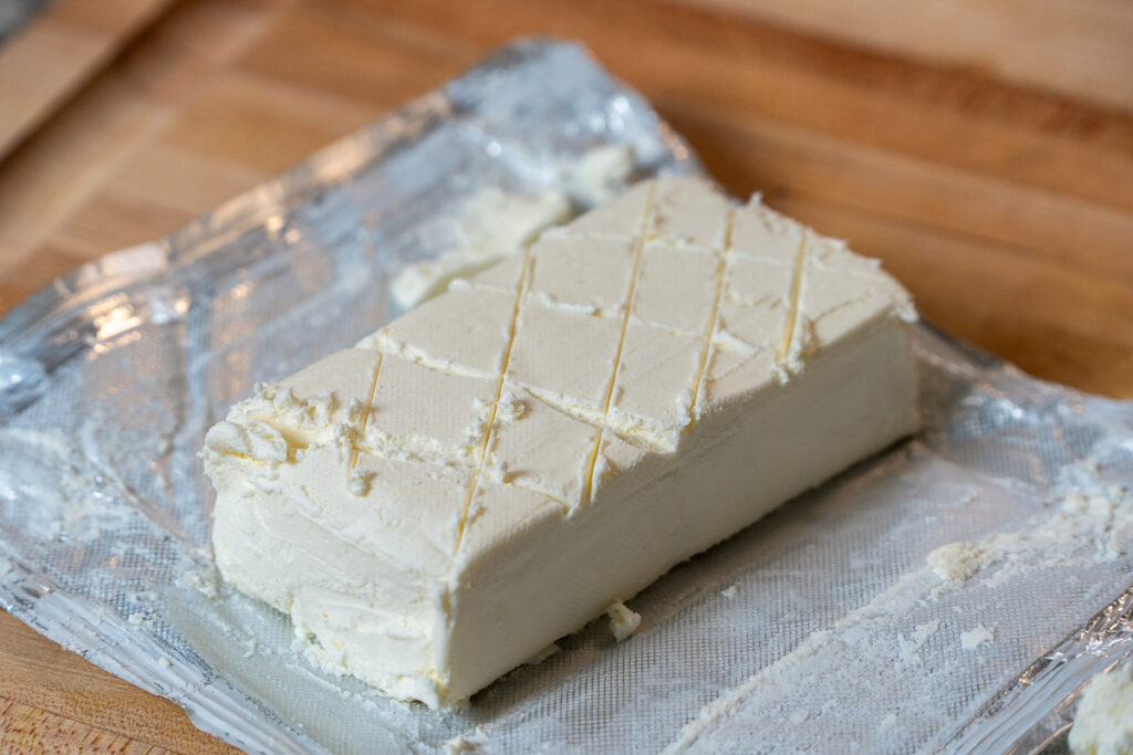 A block of cream cheese on a wooden cutting board, scored in a crosshatch pattern, with a knife beside it.