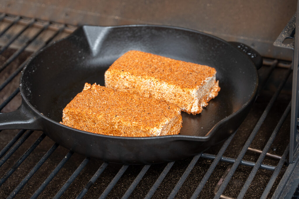 A cast iron skillet with seasoned cream cheese placed on a smoker grate at 200°F.