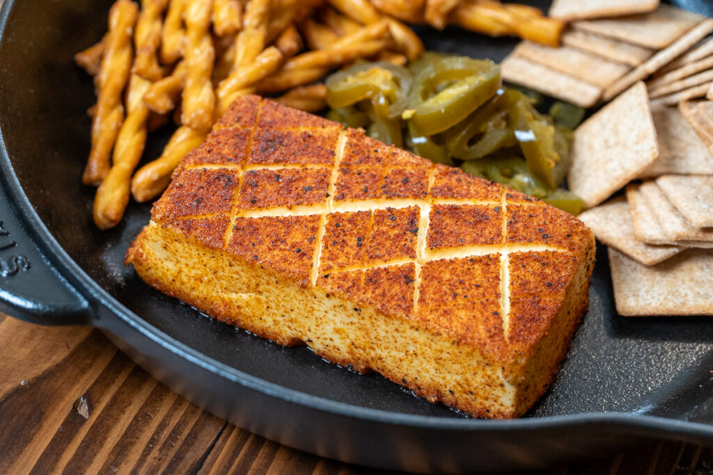 A fully smoked block of cream cheese with a golden-brown crust, served in a skillet with crackers and pickled jalapeños.