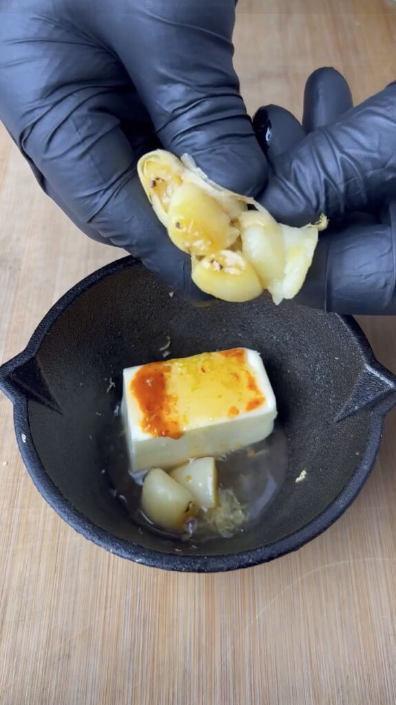 Close-up of a gloved hand squeezing roasted garlic into a small cast iron bowl with butter, lemon juice, and hot sauce for a smoky lemon garlic wing sauce.