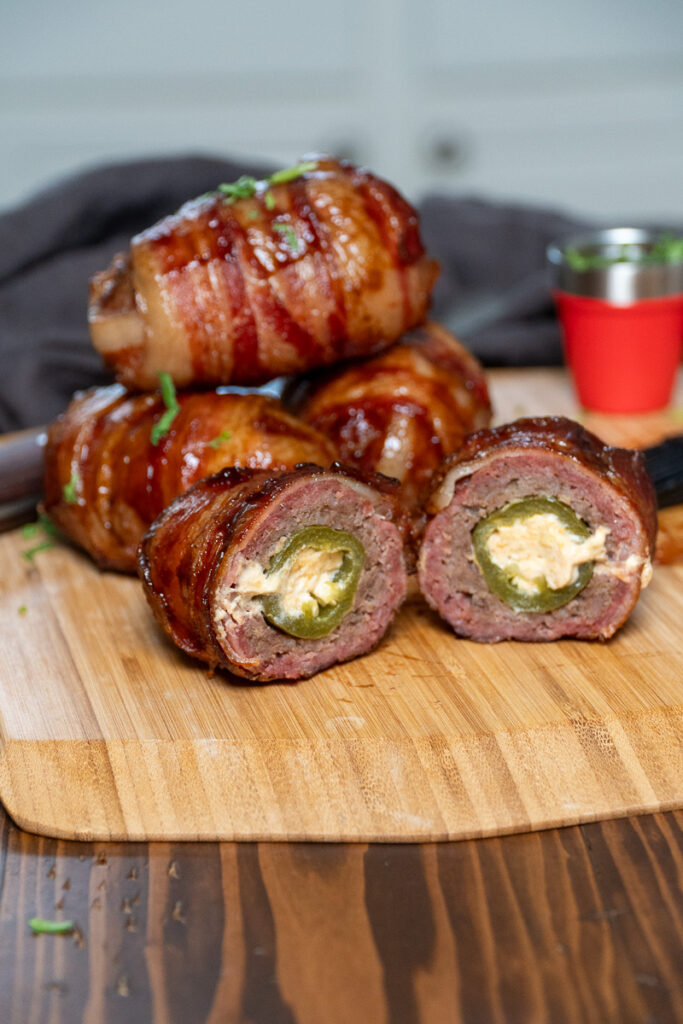 A stack of smoked Armadillo Eggs on a wooden cutting board, with one cut in half to reveal a cheese-stuffed jalapeño wrapped in sausage and crispy bacon.