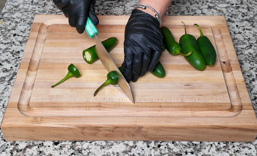 A person wearing black gloves slices the top off a jalapeño on a wooden cutting board, preparing it for stuffing. Several whole jalapeños are nearby.