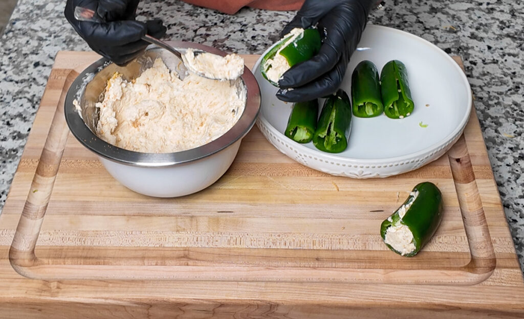 A person wearing black gloves fills a hollowed-out jalapeño with a creamy cheese mixture using a spoon. A bowl of the cheese filling and several prepared jalapeños sit nearby on a wooden cutting board.