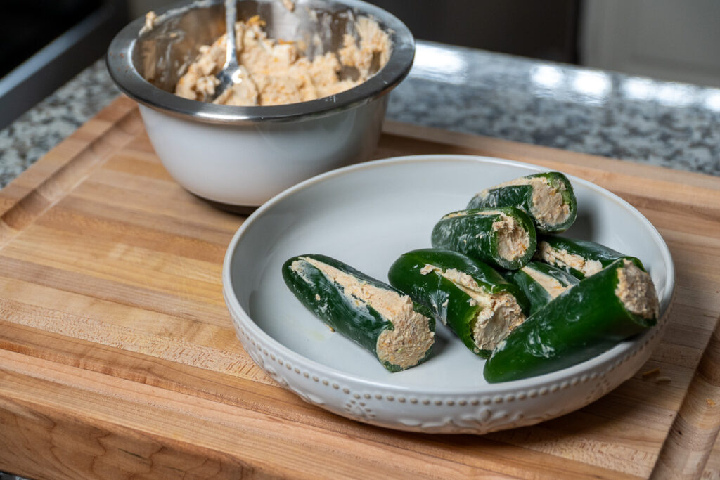 A white plate filled with jalapeños stuffed with a creamy cheese mixture sits on a wooden cutting board. A mixing bowl with leftover filling and a spoon is in the background.