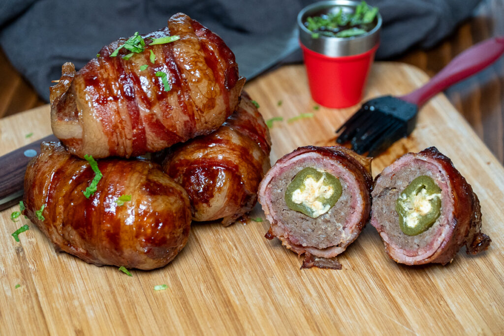 A stack of smoked Armadillo Eggs on a wooden cutting board, with one cut in half to reveal a cheese-stuffed jalapeño wrapped in sausage and crispy bacon. A brush and a cup of sauce sit in the background.