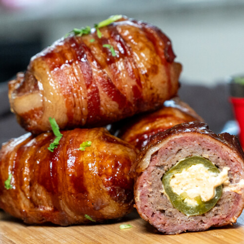 Close-up of smoked Armadillo Eggs—jalapeños stuffed with cheese, wrapped in seasoned sausage, and covered in crispy bacon, glazed with barbecue sauce and garnished with fresh herbs on a wooden cutting board.