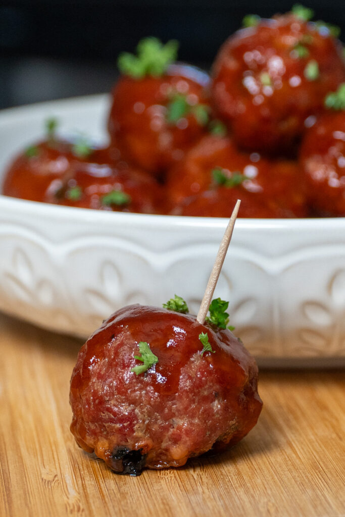 Close-up of a smoked barbecue meatball coated in a glossy BBQ glaze, garnished with fresh parsley and served with a toothpick, with a bowl of meatballs in the background.